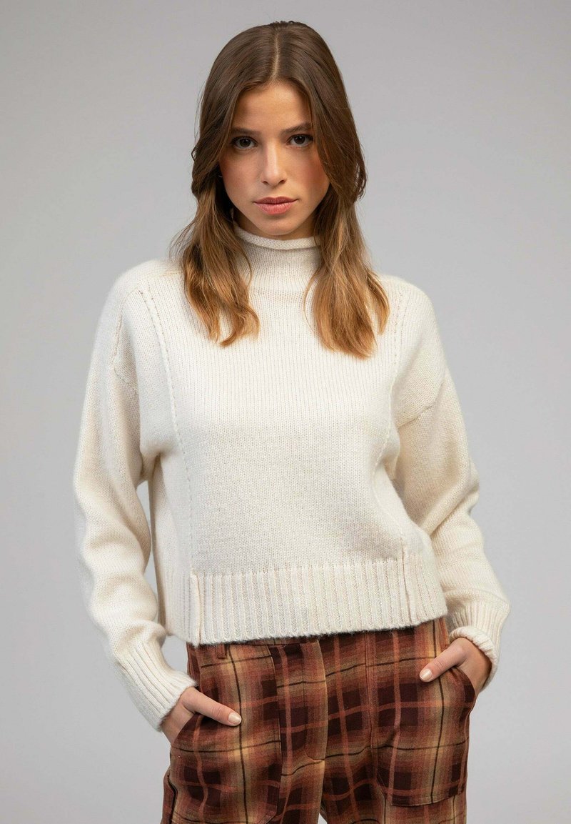 Pull en tricot blanc avec un col montant, des poignets et un ourlet côtelés. Associé avec un pantalon à carreaux marron présentant un motif à carreaux.