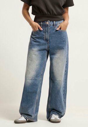 Person iført viddeblå, falmede jeans med hænderne i lommerne og hvide-grå sneakers mod en ensfarvet baggrund.