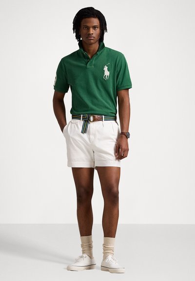 Πράσινο βαμβακερό πόλο μπλούζα με κεντημένο λογότυπο, σε συνδυασμό με λευκές βερμούδες και ριγωτή ζώνη. Το μοντέλο φορά λευκά αθλητικά παπούτσια και ανοιχτού χρώματος κάλτσες.