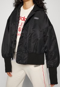 Svart satin bomberjacka med ribbat krage och manschetter. Har dragkedja och logotypdetalj på bröstet. Uppmuddiga ärmar.