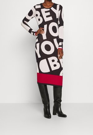 Femme portant une robe noire à manches longues avec de grandes lettres blanches et un ourlet rouge, assortie à des bottes noires à talons montant jusqu'aux genoux.