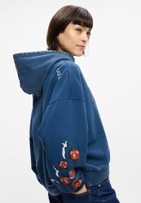 Μπλε φούτερ με κουκούλα, διακοσμημένο με κεντημένα λουλούδια σε κόκκινο και πράσινο στα μανίκια. Μαλακό ύφασμα με άνετη εφαρμογή.