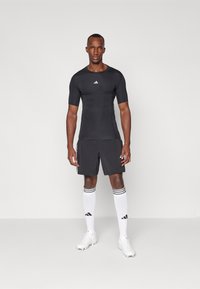 Svart atletisk skjorte med korte ermer og en hvit logo. Kombinert med svarte shorts, hvite knestrømper med svarte striper og hvite sko.