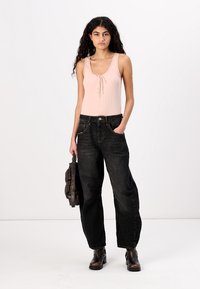 Lichtroze geribbelde tanktop met knoopdetails, gecombineerd met losse zwarte jeans en zwarte enkellaarsjes. Het model draagt een donkere tas.