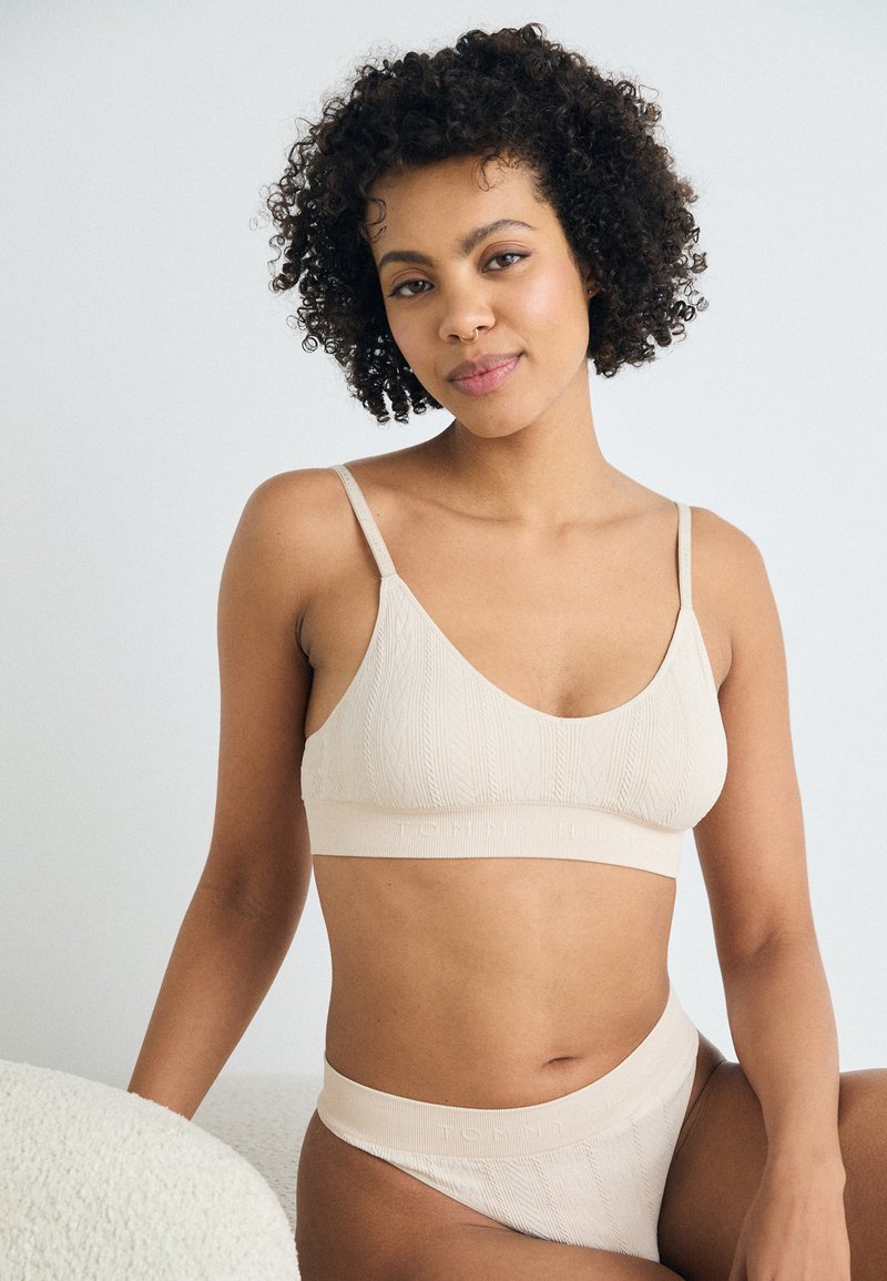 Beige niitcaga bralette õhukeste rihmadega ja sobiva kõrge vöökohaga aluspesu. Materjal on pehme, tekstuuriga ja elastne, kus on kirjas "TONI".
