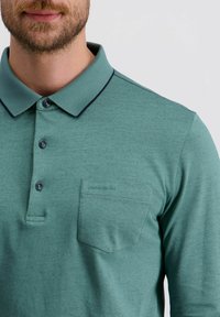 Grünes langärmliges Poloshirt aus glattem Stoff. Verfügt über eine Knopfleiste, kontrastierenden Kragenbesatz und eine Brusttasche mit Logodetail.
