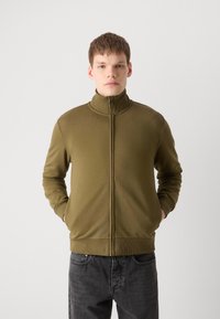 Alyvuogių žalia užtrauktuku užsegama striukė su aukštu apykakle, grublėta tekstūra ir šoninėmis kišenėmis, derinama su tamsiai pilkais džinsais.