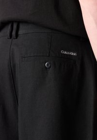 Svarte bukser med en glatt tekstur, med en baklomme, knappelukking og en liten hvit "Calvin Klein"-logo på midjebåndet.