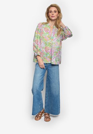 Kobieta w luźnej kwiatowej bluzce, szerokich niebieskich dżinsach i brązowych sandałach na paski, stoi z jedną ręką dotykającą włosów.