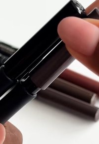 Kaksi mustaa vetoläppäistä huultenrajauskynää tyylikkäällä muotoilulla; yksi pidetään sormen välissä. Taustalla näkyy useita erilaisia ruskean sävyjä.