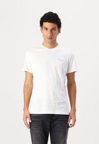 Weißes Baumwoll-T-Shirt mit Rundhalsausschnitt und kurzen Ärmeln. Verfügt über ein kleines gedrucktes Logo auf der linken Seite. Kombiniert mit dunklen Jeans.