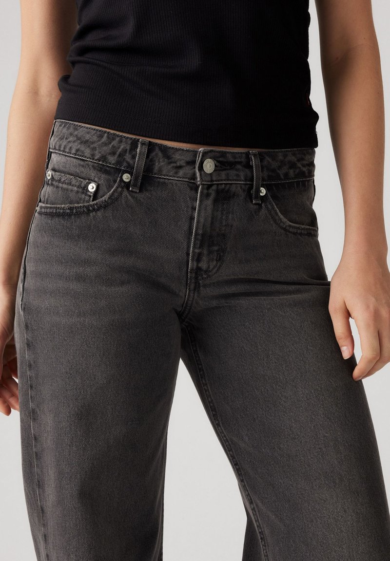 Jeans de mezclilla gris con una cintura ajustada, cinco bolsillos, un ligero desgastado y detalles en plata. El material tiene un ligero estiramiento y una textura suave.