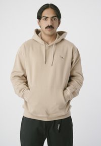 Beige Kapuzenpullover mit einer frontalen Kängurutasche, verstellbarem Kordelzug an der Kapuze und einem kleinen Vogel-Logo auf der Brust. Hergestellt aus weichem, strukturiertem Stoff.