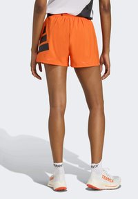 Oranžové športové šortky s čiernym bočným dizajnom, elastickým pásom a ľahkým materiálom. Nosí sa s bielymi topánkami a ponožkami.