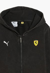 Felpa nera con zip realizzata in tessuto morbido, che presenta un logo Puma bianco e un emblema scudetto Ferrari giallo con accenti rossi e verdi sulla tasca.