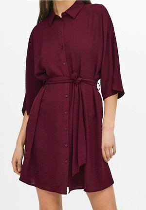 Femme portant une robe boutonnée bordeaux foncé, longueur genou, manches trois-quarts et ceinture nouée à la taille.