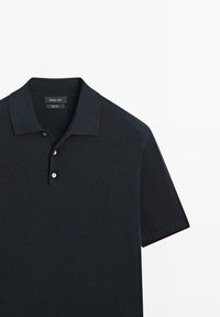 Sort kortærmet polo med krave og tre hvide knapper, lagt fladt på en hvid baggrund.