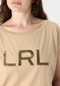 Beige kortärmad t-shirt med metalliskt gyllene broderade bokstäver "LRL" på framsidan, med bred ringning och mjuk bomullstextur.