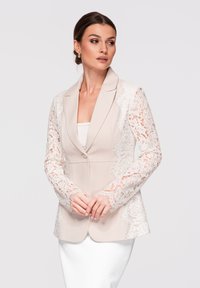 Femme portant un blazer beige avec des manches en dentelle blanche délicate et des panneaux latéraux, associé à une robe blanche, regardant sur le côté.