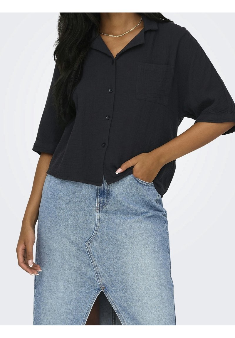 Personne portant une chemise noire boutonnée avec une poche poitrine et une jupe en jean bleu clair avec une fente à l'avant, main dans la poche de la jupe.