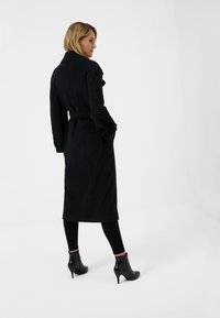 Svart bältejacka i mjukt tyg, med krage och uppvikta ärmar, tillsammans med tajta svarta leggings och ankelboots.