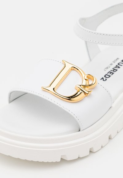 Sandalo platform bianco con una larga fascia caratterizzata da un logo "D" dorato. Materiale liscio, suola spessa con dettagli testurizzati, design minimalista.