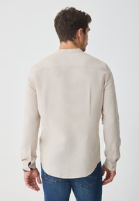 Beige langærmet skjorte lavet af let stof, med en afslappet pasform, rygskyd og knapmanchetter. Bæres med blå jeans.