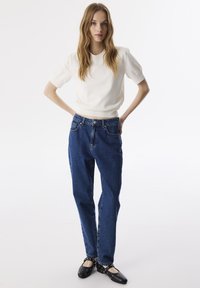 Weißer Pulli mit kurzen Ärmeln, kombiniert mit hochgeschnittenen blauen Denim-Jeans. Schwarze Ballerinas mit Nieten-Details runden den Look ab.