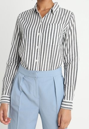Woman wearing a gray and white vertical striped button-up shirt tucked into high-waisted light blue pleated pants.