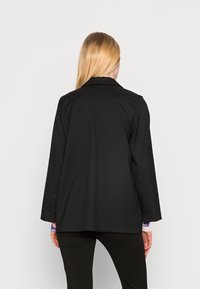 Veste noire cintrée avec col, manches longues et coupe décontractée, présentant une texture lisse et un design minimaliste.