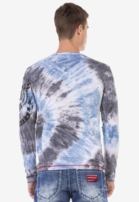 Langärmliges Batikshirt mit blauen, grauen und weißen Wirbeln. Verfügt über ein schwarzes Grafikdesign auf dem linken Ärmel und ein rotes Etikett am Saum.