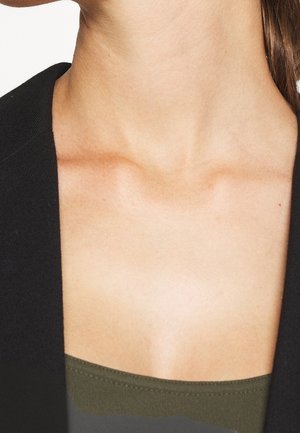 Gros plan sur le cou et le haut de la poitrine d'une femme portant un blazer noir sur un haut vert olive.