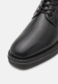 Chaussure en cuir noir avec un bout rond, texture lisse et semelle en caoutchouc épaisse. Présente des lacets noirs et des coutures le long du bord de la semelle.