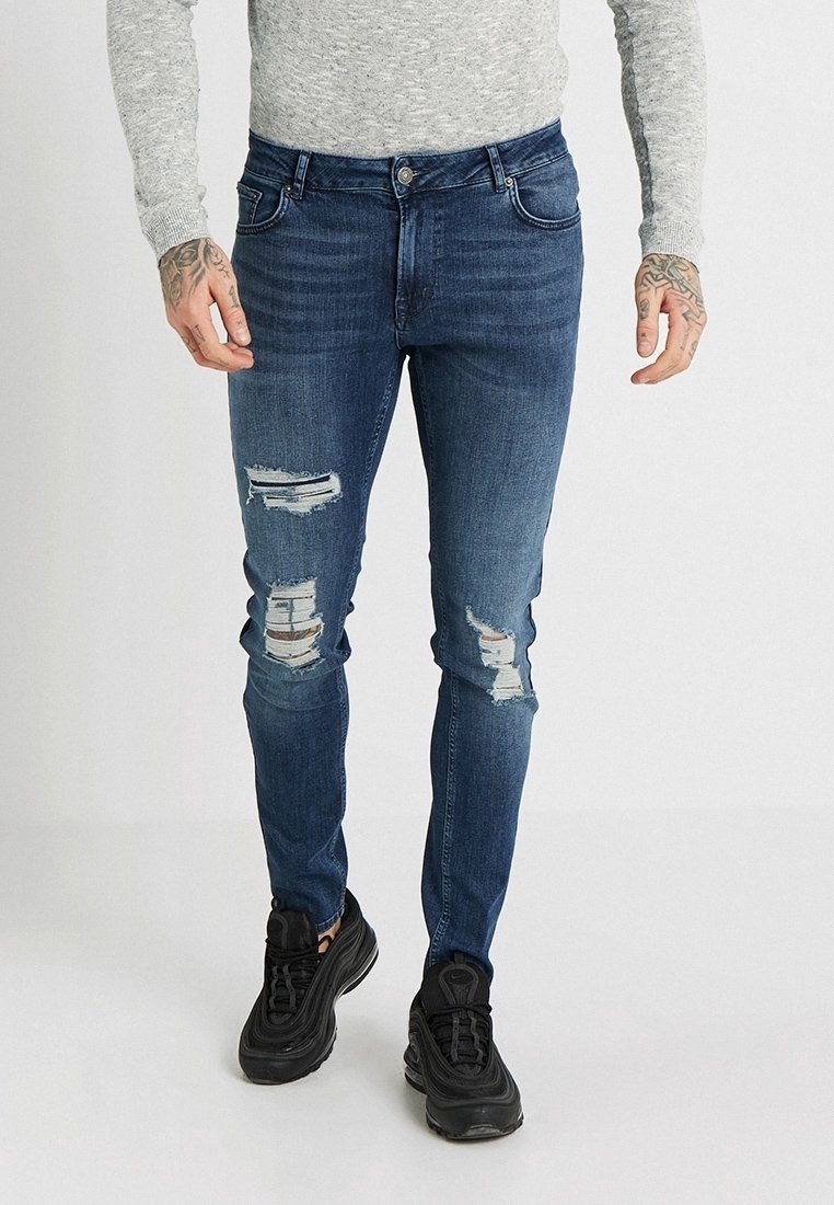 Jeans de mezclilla de un azul oscuro desgastado, con múltiples rasgaduras, corte ajustado y un sutil desgastado, combinados con zapatillas deportivas negras.