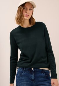 Femme aux cheveux châtain clair mi-longs portant une casquette beige, un pull à manches longues vert foncé et un jean bleu sur un fond clair.