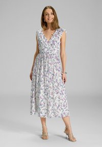 Frau steht und lächelt in einem ärmellosen weißen Midikleid mit floralem Muster und Rüsche an der Schulter, trägt sandalen mit hohen Absatz und Riemchen.