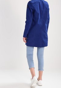 Lång blå kappa i mjukt tyg, med en skräddarsydd silhuett och en ryggslits. Bärs över ljusblå uppvikta jeans och vita sneakers.