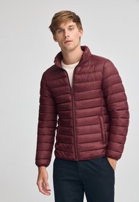 Burgundy puffjacka med dragkedja fram, ståkrage och horisontell quiltad design. Har en liten logotyp på vänster bröst.