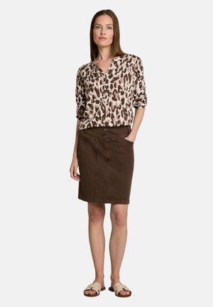 Frau, die eine braune Bluse mit Leopardenmuster, einen braunen knielangen Rock und beigefarbene Sandalen mit offenen Zehen trägt, steht mit einer Hand in der Tasche.