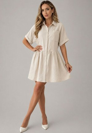 Femme portant une robe beige à manches courtes boutonnée avec une poche avant, debout avec une main sur la hanche et des talons blancs.