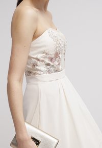 Hihaton kerma-coloured mekko, jossa on kukkakirjontaa, levenevä hameosa ja satiinivyötärö. Malli pitää käsissään teksturoitua kultaklutchia.