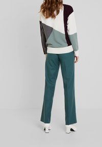 Vrouw met lang haar in een veelkleurige geometrische trui, teal broek en witte blokhak enkellaarsjes, staande tegen een grijze achtergrond.