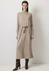 Béžové pletené šaty s vysokým krkom, dlhými rukávmi a stiahnutým pásom s hnedým opaskom, kombinované s hnedými čižmami po stehná. Hladká textúra.