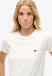 Biała bawełniana koszulka z okrągłym dekoltem i krótkimi rękawami, z małym czarnym logo "Super Dry" na lewej stronie klatki piersiowej.