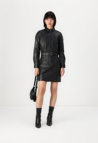 Čierne kožené šaty vo fitted siluete s špicatým golierom a opaskom v páse. Modelka nesie malú čiernu kabelku a má na sebe čierne kotníkové topánky.