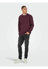 Bordeaux sweatshirt met een ronde hals, gecombineerd met donkere jeans en beige sneakers. De stof lijkt glad en heeft een lichte textuur.