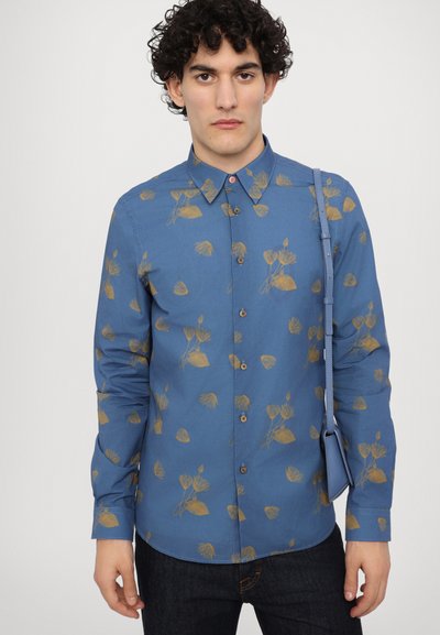 Camisa azul de botones con un patrón floral dorado, de manga larga y con cuello. Combinada con jeans de mezclilla oscuros y un bolso azul de hombro.