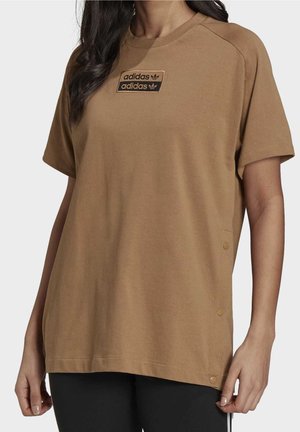 Kobieta nosząca luźną brązową koszulkę Adidas z podwójną naszywką z logo na piersi i zatrzaskami wzdłuż bocznego szwu.