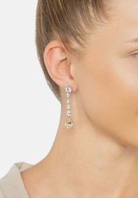 Egy nő jobb füle hosszú, lógó fülbevalót visel, amelyen egy nagy és öt kisebb átlátszó és világos rózsaszín drágakő található.