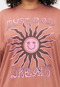 Camiseta de gran tamaño marrón con un gráfico de sol rosa que tiene una cara sonriente y el texto "Sueños Místicos" en una tipografía caprichosa.